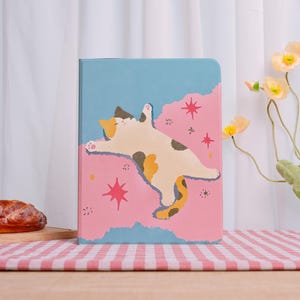 Peut inclure: Un carnet avec une illustration fantaisiste d'un chat détendu. La couverture présente un fond bleu clair et rose avec un motif ludique d'un chat dans une pose détendue, agrémenté d'éléments en forme d'étoile. Le chat est blanc, marron et jaune.
