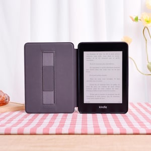 Pode incluir: Um leitor de e-books Kindle preto com uma capa cinza, aberto para exibir texto. A capa tem uma al&ccedil;a para segurar o dispositivo. A tela mostra texto em franc&ecirc;s. Em uma superf&iacute;cie xadrez rosa e branca.