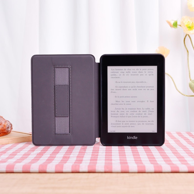 Pode incluir: Um leitor de e-books Kindle preto com uma capa cinza, aberto para exibir texto. A capa tem uma al&ccedil;a para segurar o dispositivo. A tela mostra texto em franc&ecirc;s. Em uma superf&iacute;cie xadrez rosa e branca.