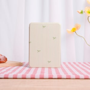 May include: A rectangular, off-white case with vertical stripes and small green bow patterns. The case is set on a pink and white checkered tablecloth, with a wooden surface below. A flower and white curtain are in the background.