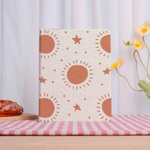 May include: A beige notebook with a repeating pattern of orange suns, stars, and dots. The notebook is standing upright on a wooden surface with a red and white checkered tablecloth. Decorative flowers are in the background.