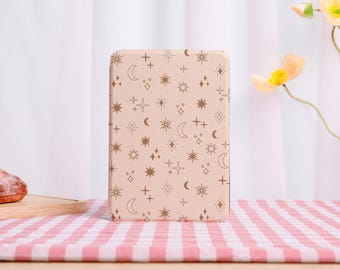 May include: A rectangular, light pink box with rounded corners. The box is covered in a repeating pattern of brown stars, moons, and crosses. The box is sitting on a red and white checkered tablecloth.