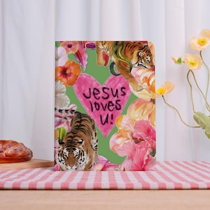 Peut inclure: Un journal avec un fond vert, représentant un cœur rose avec le texte "Jesus loves U!" et des illustrations de tigres et de fleurs colorées. Le journal est sur une table avec une nappe à carreaux rouges et blancs.