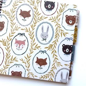 May include: A spiral-bound notebook featuring a white cover with a repeating pattern of gold leafy branches and oval frames containing illustrations of foxes, bears, and rabbits. Four side tabs are labeled Our Journey, Waiting for You, We're Matched, and Welcome Home.