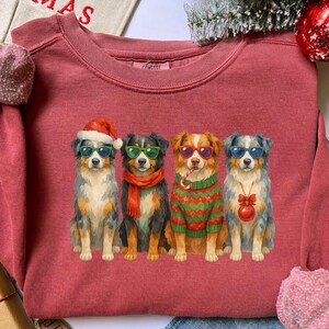 May include: A red-toned sweatshirt featuring a graphic of four dogs wearing sunglasses and festive holiday attire. One dog wears a Santa hat, another a scarf, a third a sweater, and the last a Christmas ornament. The word "MERRY CHRISTMAS" is visible.