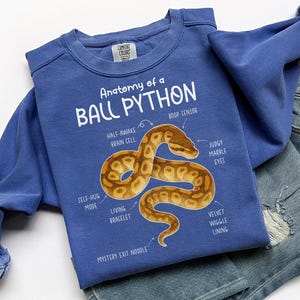 May include: A blue sweatshirt featuring the text "Anatomy of a Ball Python" and a graphic of a brown and tan snake. The sweatshirt also has the words "Half-Awake Brain Cell", "Boop Sensor", "Self-Hug Mode", "Living Bracelet", "Mystery Exit Noodle", "Judgy Marble Eyes", and "Velvet Wiggle Lining".