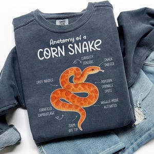 Op de afbeelding: Een marineblauw sweatshirt met de tekst "Anatomy of a Corn Snake" en een illustratie van een oranje slang. De slang heeft labels die naar zijn kenmerken verwijzen, waaronder "Spicy Noodle" en "Zoom-Off Tip."