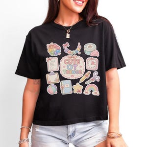May include: A black t-shirt featuring a central graphic with the text "CUTE OT THINGS" surrounded by pastel-colored illustrations of a pop-it toy, beads, putty, a clipboard, a timer, a pencil, scissors, a ball, a notebook, a star, and a rainbow.