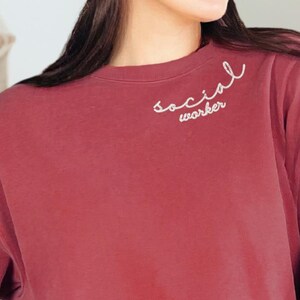 May include: A maroon sweatshirt with the words "social worker" embroidered in white script on the upper left chest. The long-sleeved crew neck shirt is paired with blue jeans.