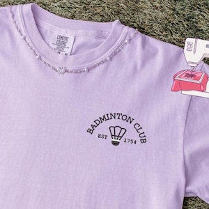 Może przedstawiać: Jasnofioletowa koszulka z napisem "Badminton Club" i grafiką lotki. Koszulka ma postrzępiony dekolt i napis "EST 1754". Koszulka wykonana jest z miękkiego materiału.