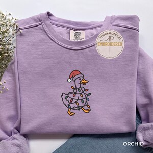 Peut inclure: Sweat-shirt lilas avec un motif brodé d'un canard portant un bonnet de Père Noël et des guirlandes lumineuses. Le sweat-shirt à col rond a des manches longues. L'étiquette indique "Comfort Colors". Un autocollant en haut à droite indique "Personalized Gift Embroidered".