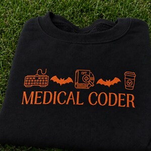 Puede incluir: Una sudadera negra con bordado naranja. El diseño incluye un teclado de ordenador, un murciélago, un libro con la etiqueta CPT y una cruz médica, otro murciélago y un vaso de café. El texto MEDICAL CODER está bordado en naranja debajo de los iconos.