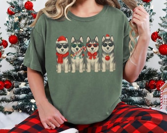 May include: An olive green t-shirt featuring a graphic of five cartoon huskies wearing sunglasses and holiday accessories. The dogs are dressed in a Santa hat, scarf, string lights, bow tie, and ornament. The person in the image is wearing red and black plaid pajama pants.