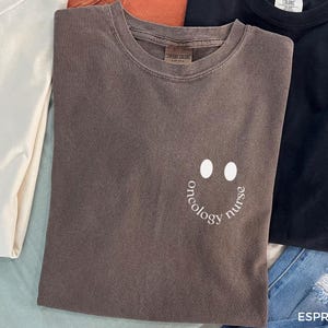 May include: A folded espresso-coloured t-shirt with a smiley face design made of two white ovals and the words "oncology nurse" in a curved font. Other folded t-shirts in cream, orange, and black are visible.