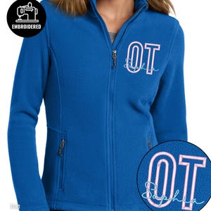 May include: A royal blue fleece zip-up jacket featuring a stand-up collar and two side pockets. The left chest is embroidered with the letters OT in a light pink outline font, with the name Sophia written in a light blue script below.