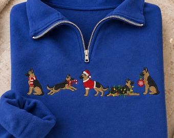 May include: A royal blue quarter-zip sweatshirt featuring an embroidered row of five German Shepherd dogs. The dogs are depicted wearing a red and white striped scarf, holding a candy cane, wearing a Santa suit, tangled in lights, and holding an ornament.