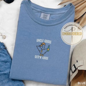 以下が含まれることがあります： 「UNCLE GOOSE GETT'N LOOSE」の刺繍とボトルを持った漫画のガチョウが描かれた、ブルーデニムカラーのTシャツ。クルーネックで、サングラス、ネックレス、サンダルなどのアクセサリーに囲まれています。