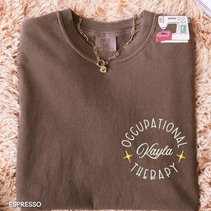 May include: Espresso-colored Comfort Colors t-shirt with a gold chain necklace. The shirt features embroidered text that reads "Occupational Therapy Kayla" in a circular design. A small sticker of a sewing machine is visible.