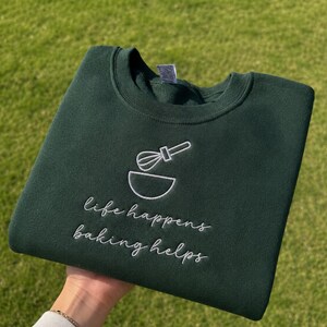 May include: A dark forest green crewneck sweatshirt featuring white embroidery of a whisk and a mixing bowl. Below the graphic, the text reads "life happens baking helps" in a white cursive script. The garment has a ribbed collar and cuffs.