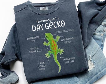 Puede incluir: Una sudadera azul marino con el texto "Anatomy of a Day Gecko" y una ilustración de un gecko verde. El gecko tiene manchas naranjas y términos anatómicos humorísticos. La sudadera está doblada sobre unos vaqueros azules.