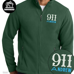 May include: A forest green full-zip fleece jacket featuring a high collar and long sleeves. The chest displays white embroidered text reading 911 with the name ANDREW embroidered in light blue underneath. A circular badge indicates it is embroidered.