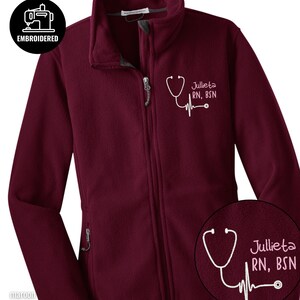 Puede incluir: Chaqueta de forro polar color granate con cremallera completa y dos bolsillos laterales. La chaqueta presenta un diseño de estetoscopio blanco bordado con el nombre "Jullieta RN, BSN". La chaqueta tiene un cuello alto y la palabra "bordado" está impresa en un parche circular.