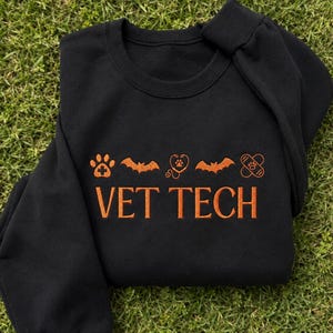 May include: A black long-sleeve sweatshirt featuring orange embroidery. The design includes a row of icons: a paw print with a cross, a bat, a heart-shaped stethoscope with a paw print, another bat, and a bandage. Below, the text reads VET TECH.