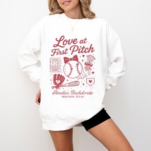 Peut inclure: Sweat-shirt blanc avec un motif rouge sur le thème du baseball. Le motif comprend le texte "Love at First Pitch", une balle de baseball, un nœud, un gant et d'autres éléments. Le texte comprend également "Amelia's Bachelorette" et "Houston, Texas."