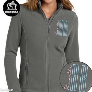 May include: A deep smoke gray fleece jacket with a full zipper closure and two side pockets. The jacket features the embroidered text "EMBROIDERED" in a black circle. The initials "HR" are embroidered in blue and pink, with the name "Samantha" in pink.