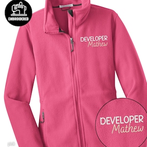 Può includere: Giacca in pile rosa con cerniera intera e due tasche laterali. La giacca presenta la scritta ricamata "DEVELOPER Mathew" in bianco sul petto. Nell'angolo in alto a sinistra è presente un logo circolare con la scritta "EMBROIDERED" e l'icona di una macchina da cucire.