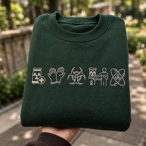 Op de afbeelding: Een donker bosgroene sweater met ronde hals en een reeks van vijf geborduurde witte lijntekeningen op de borst, waaronder een radioactieve container, beschermende handschoenen, een biohazard-symbool, een werkend persoon en een atoom.