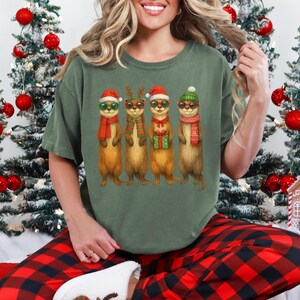 May include: Green t-shirt with a Christmas otter design. The otters are depicted wearing Santa hats, sunglasses, and scarves, with one holding a gift. The model is wearing red and black plaid pajama bottoms.