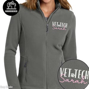 May include: A deep smoke gray fleece jacket with a full zipper closure and two side pockets. The jacket is embroidered with the words "VET TECH Sarah" in white and pink lettering. The jacket also has an embroidered patch that says "EMBROIDERED".