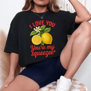 May include: Black t-shirt with a lemon graphic and the text "I LOVE YOU, You're my squeezee" in red. The graphic includes a whole lemon, a lemon slice, leaves, and white flowers. The shirt is paired with navy blue shorts.