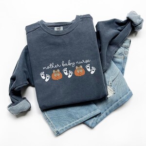 May include: A navy blue sweatshirt with the text "mother baby nurse" and a graphic of two pumpkins and baby footprints. The sweatshirt is folded over a pair of light blue denim jeans.
