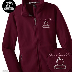 May include: Maroon fleece jacket with a full zipper and a high collar. The jacket features the embroidered text "Mrs. Smith" above a white apple and book design. A small circular graphic in the upper left corner reads "EMBROIDERED".