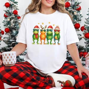 May include: White t-shirt with a watercolour illustration of four frogs in sunglasses and festive holiday outfits. The frogs are green and orange, with Santa hats, scarves, and reindeer antlers. The shirt is worn with red and black plaid pyjama bottoms.