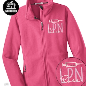 May include: A pink fleece jacket with a full zipper closure. The jacket features an embroidered design of a syringe, the letters "LPN", and the name "Rose" in white. The jacket also has two side pockets with zippers and the word "EMBROIDERED" in a black circle.