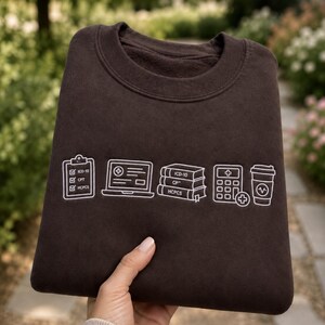 May include: A dark brown crewneck sweatshirt featuring white embroidered line art icons of a medical clipboard, a laptop, a stack of three books, a calculator with a plus sign, and a coffee cup, all labelled with ICD-10, CPT, and HCPCS text.