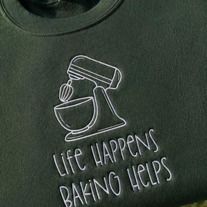 May include: A dark forest green crewneck sweatshirt featuring white embroidery of a stand mixer with a mixing bowl. Below the mixer, the text "LIFE HAPPENS BAKING HELPS" is embroidered in white, using a simple, clean, and legible sans-serif font.