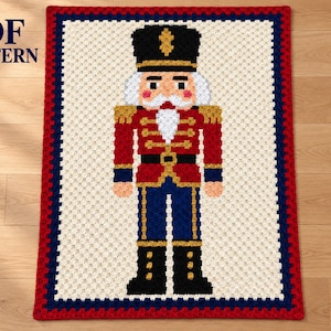 May include: A rectangular crochet blanket featuring a nutcracker design with a black hat, red jacket, blue trousers, and black boots. The central figure is set against a cream background with a dark blue and red border. Text reads PDF PATTERN.