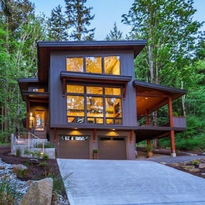 Op de afbeelding: Modern huis met twee verdiepingen, een grijze buitenkant, grote ramen en een bruin dak. Het huis heeft een garage voor twee auto's en een betonnen oprit, omringd door groene bomen.