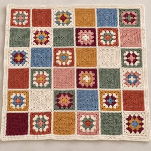 May include: A square crochet blanket composed of a grid of multi-colored granny squares and solid cream-colored squares. The pattern features shades of terracotta, mustard yellow, sage green, dusty blue, and deep burgundy yarn.