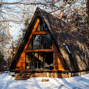 May include: A-frame cabin with a dark roof and wooden siding, nestled in a snowy forest. Large windows offer views of the surrounding trees. The cabin sits on a foundation of stacked logs, with a wooden staircase leading up to the entrance.