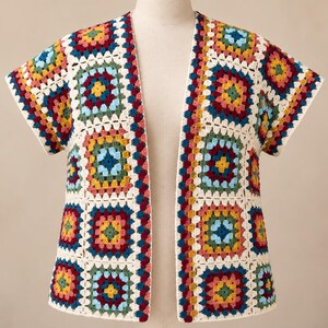 May include: A short-sleeved cardigan constructed from multi-colored granny square crochet motifs. The garment features a repeating pattern of red, yellow, blue, and green yarn squares joined by white borders, with an open front and no buttons.
