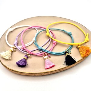 Puede incluir: Una colección de pulseras con borlas de colores blanco, rosa, morado, azul, amarillo y naranja. Cada pulsera presenta un dije de borla y se exhibe sobre un plato redondo de tono neutro.