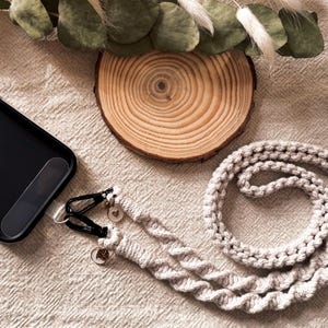May include: A black smartphone case with a light gray woven phone strap. The strap has a braided design and is attached to the case with black clasps and small charms. The background includes a wooden slice and decorative greenery.