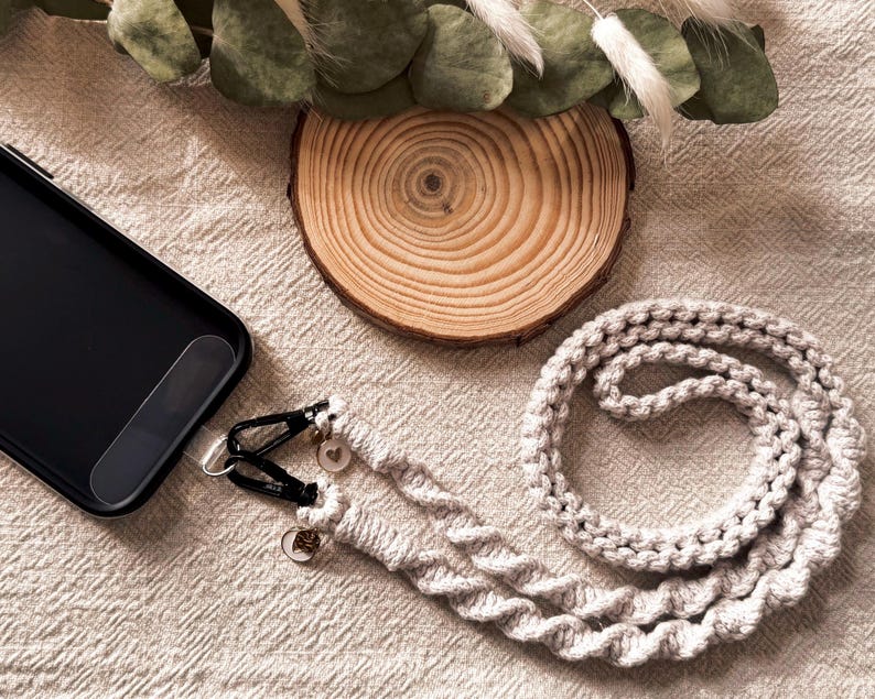 May include: A black smartphone case with a light gray woven phone strap. The strap has a braided design and is attached to the case with black clasps and small charms. The background includes a wooden slice and decorative greenery.
