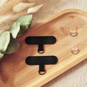 May include: An overhead shot displays a wooden tray holding several phone grip accessories. Two black, rectangular grip tabs with silver rings are positioned near two clear, rectangular tabs. Dried plants and greenery are arranged on the left side of the tray.
