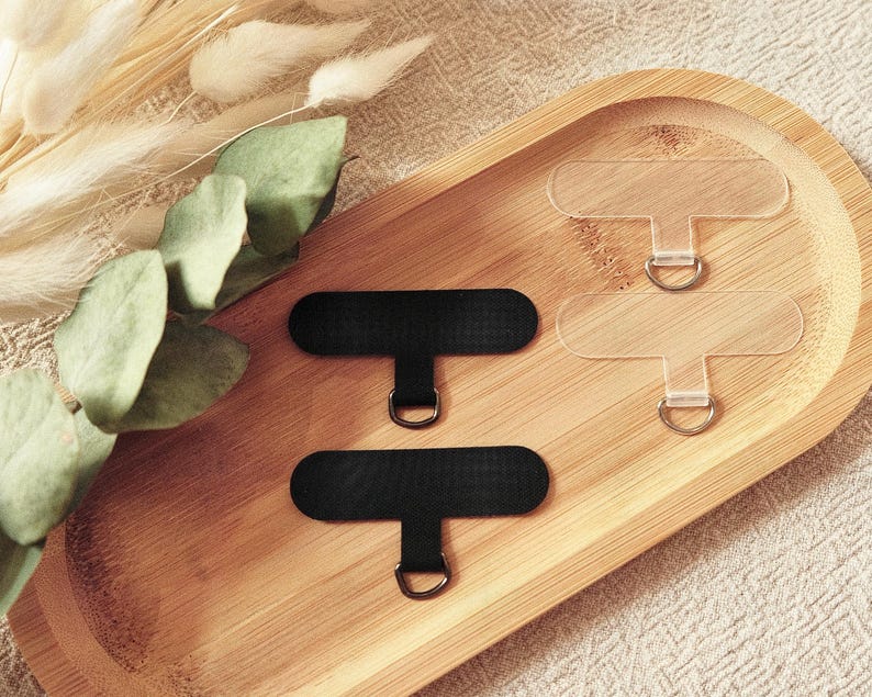 May include: An overhead shot displays a wooden tray holding several phone grip accessories. Two black, rectangular grip tabs with silver rings are positioned near two clear, rectangular tabs. Dried plants and greenery are arranged on the left side of the tray.